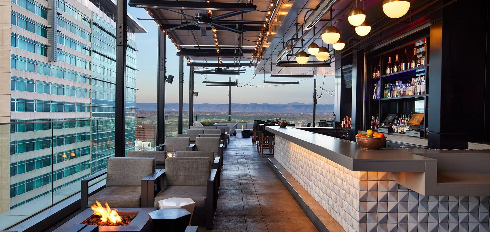 A glass-walled rooftop bar with a fire pit, lounge seating and a lit counter, with city towers outside.