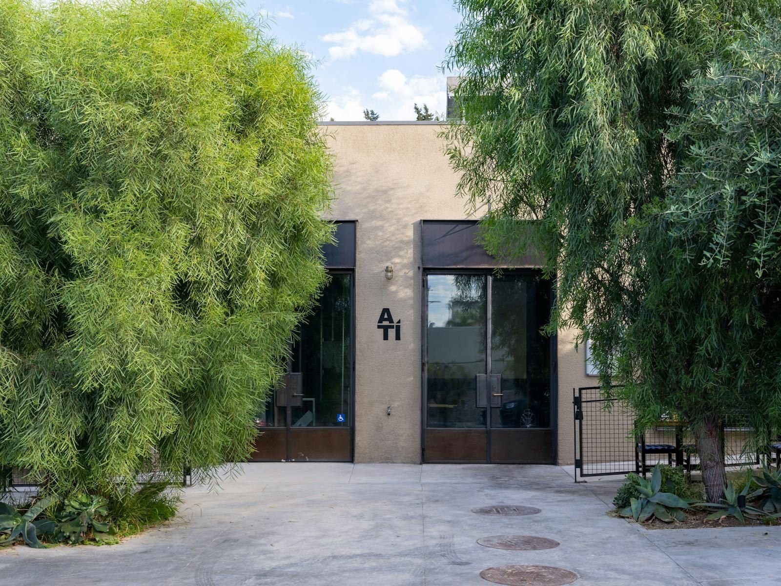 Entrance to A Tí set behind tall green trees, with a paved courtyard in front of the glass doors