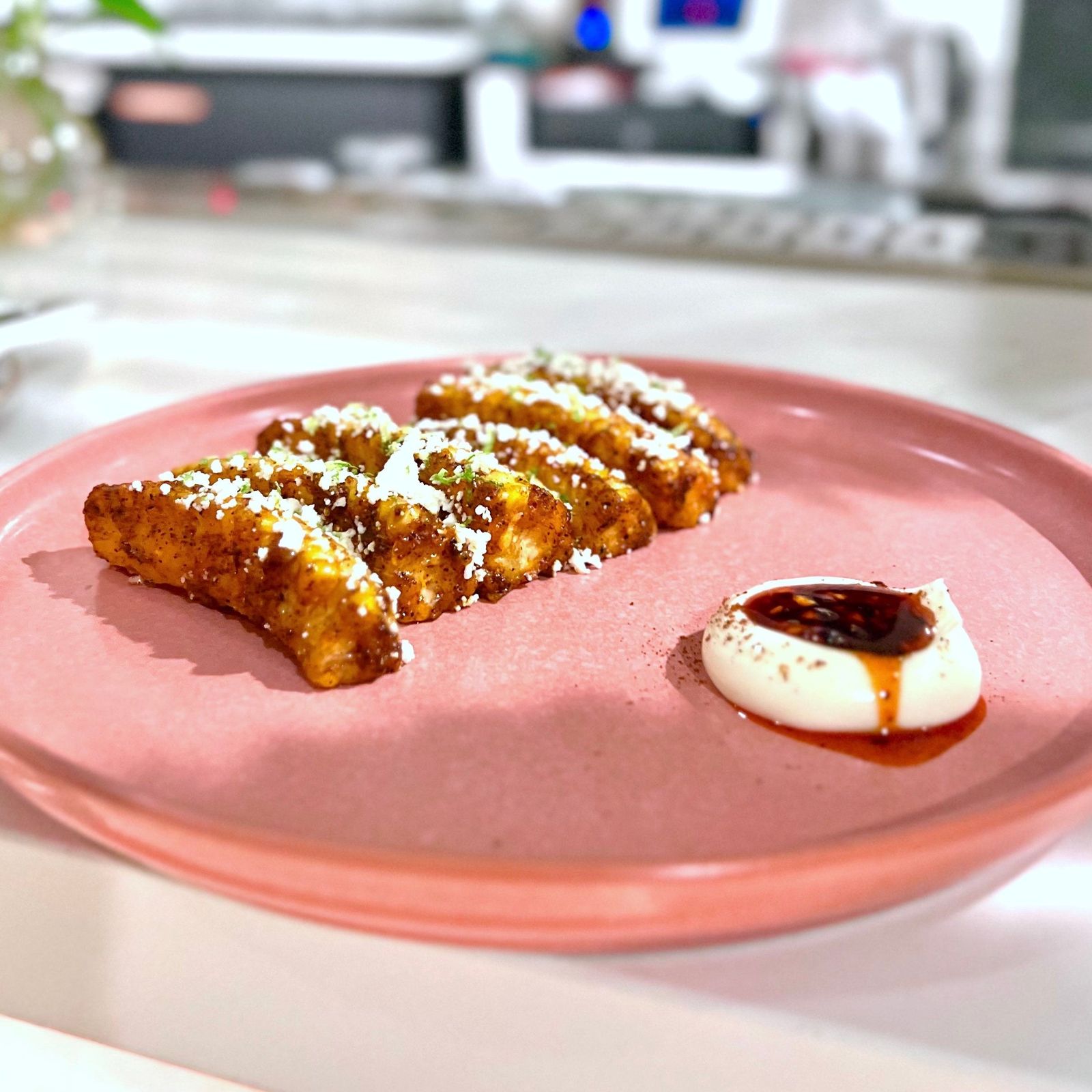 Fried plantain pieces on a pink plate with a spoonful of dark sauce and cream