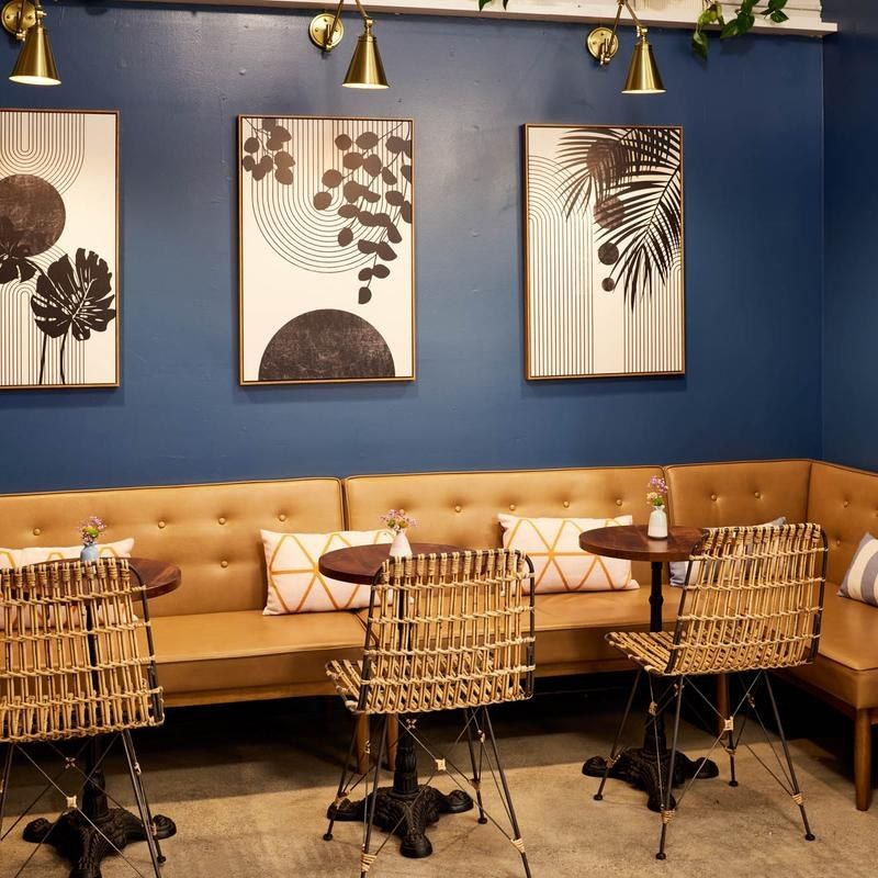 A dining nook with a tan leather banquette, wicker chairs and framed botanical prints on a deep blue wall.