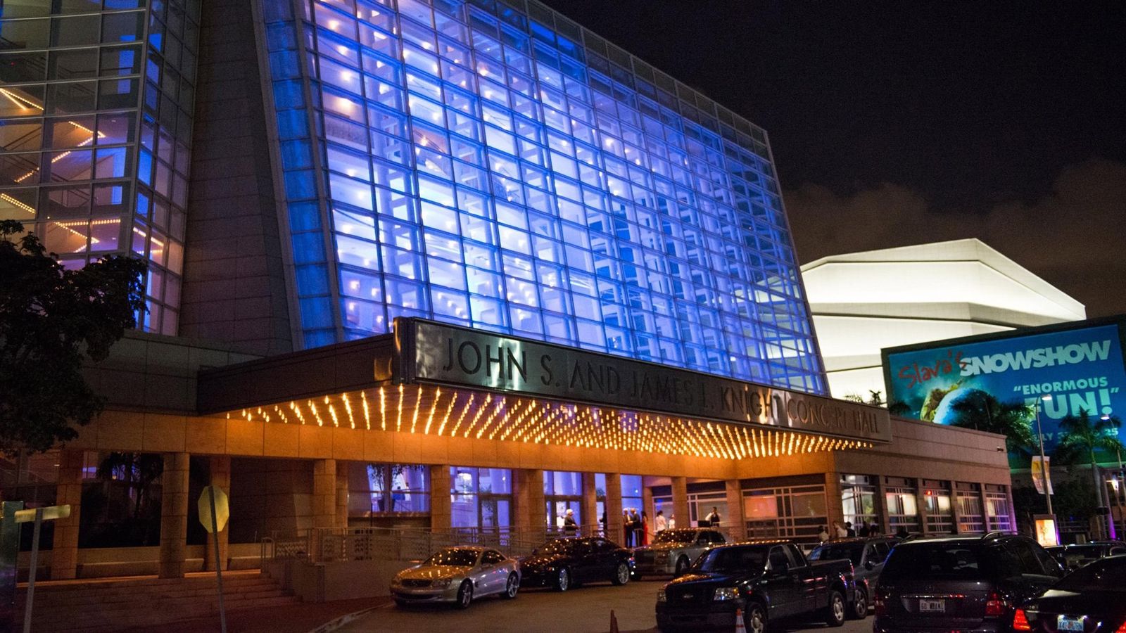 The glass facade of the Adrienne Arsht Center lit blue at night above a covered entrance