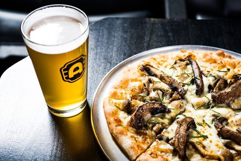 A pint of beer beside a mushroom pizza on a dark table.
