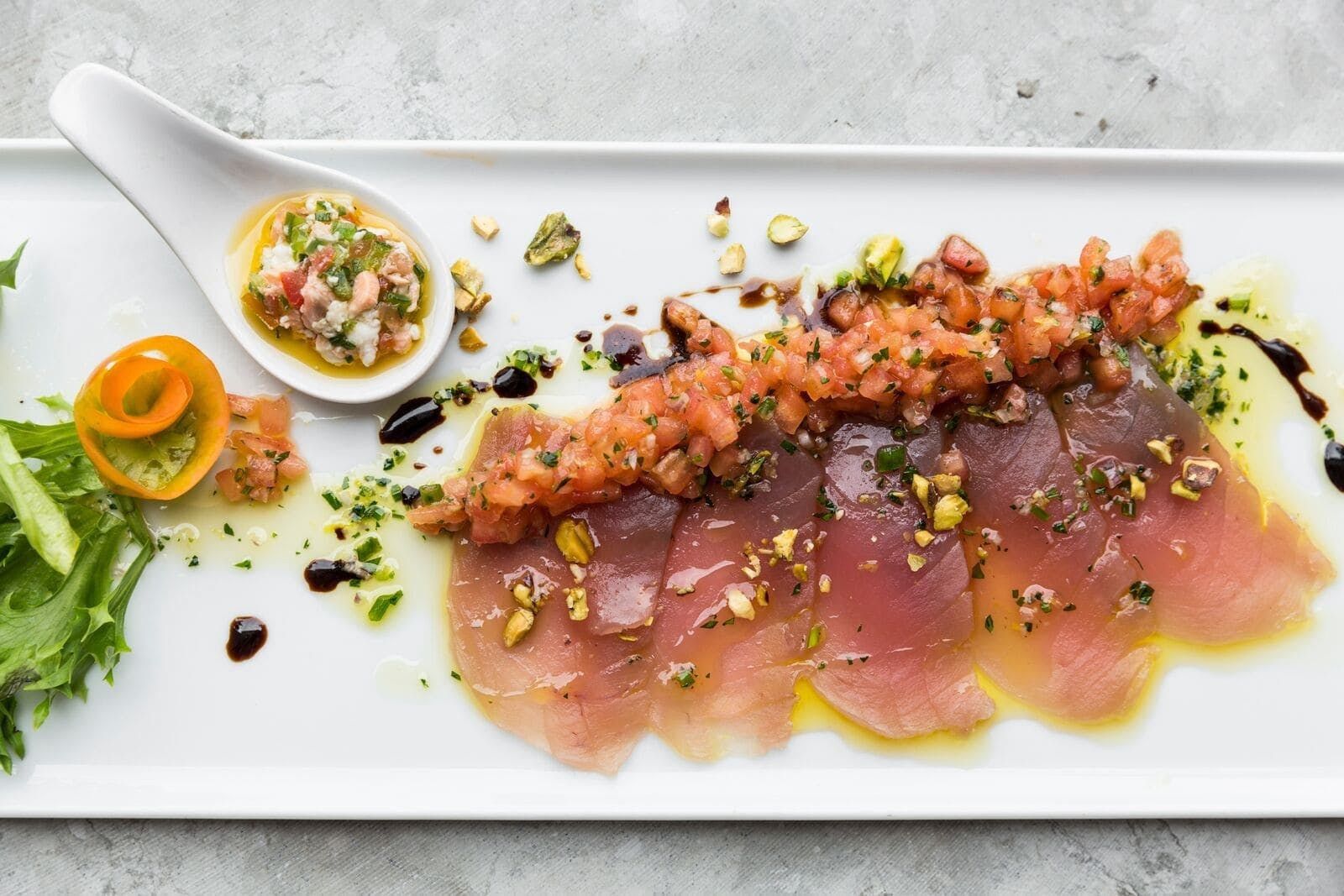 Sliced raw tuna topped with diced tomato on a long white plate with a spoon of garnish