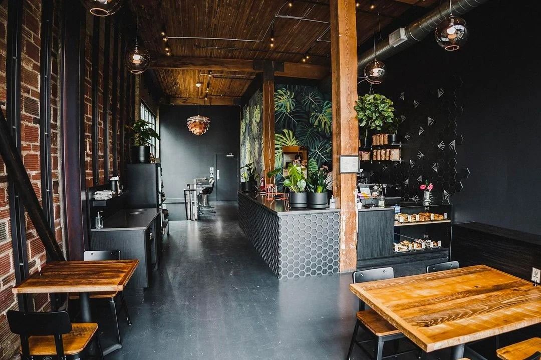 A long cafe room with a brick wall, wood beams, hanging lamps, a hexagon-tiled counter and wooden tables.