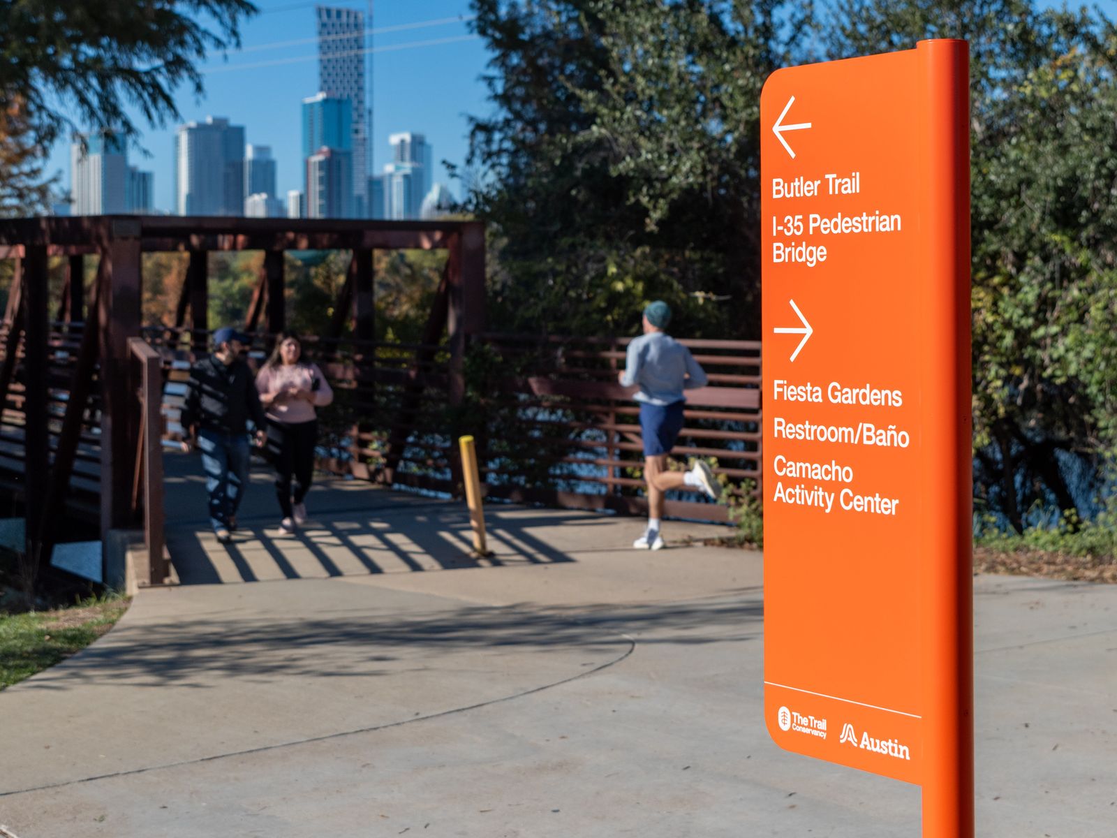 Runners on a paved trail beside an orange trail sign, with a wooden bridge and the Austin skyline beyond.