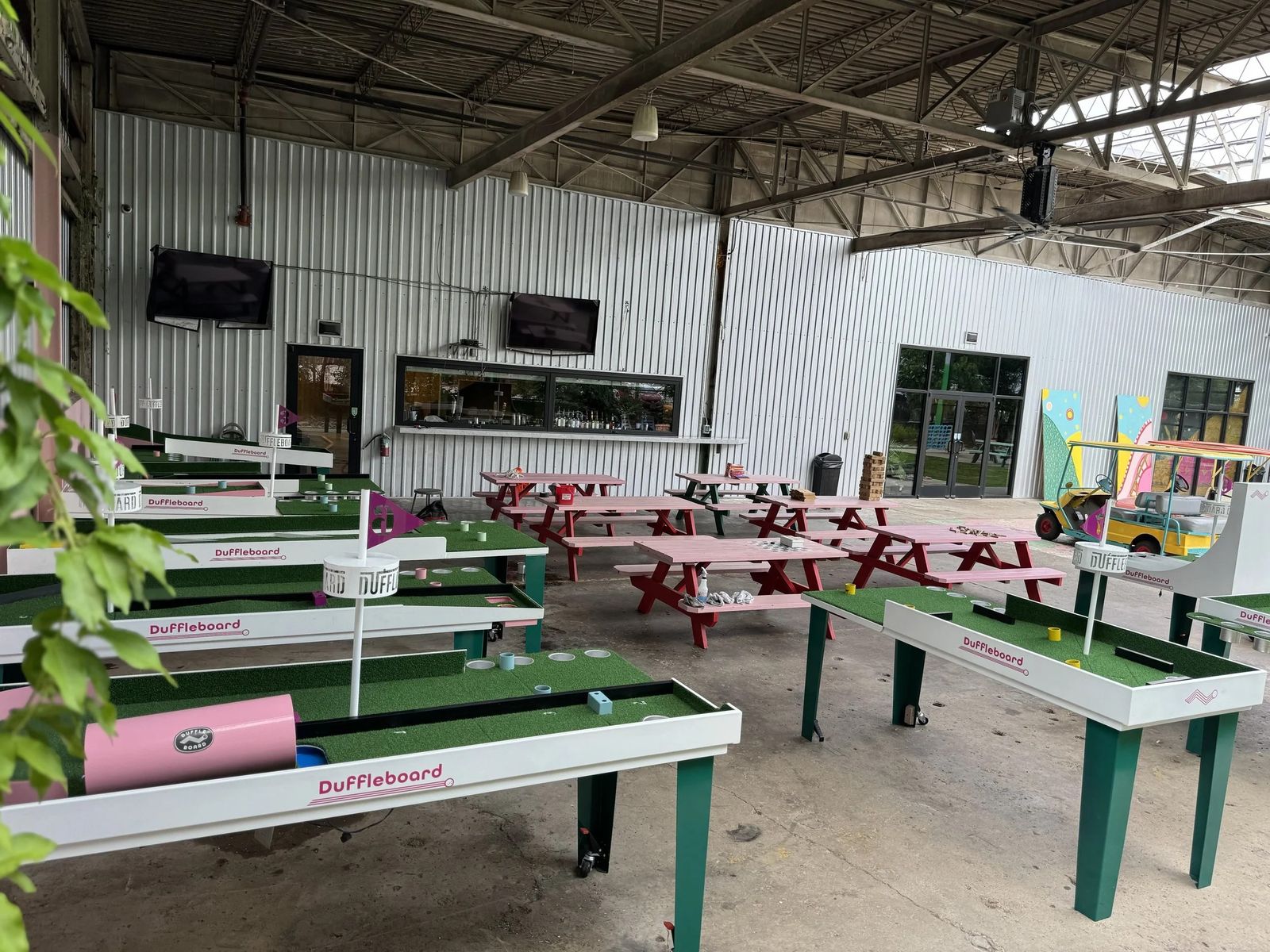 A large warehouse space with rows of indoor mini-golf holes and shuffleboard-style tables under a metal roof.