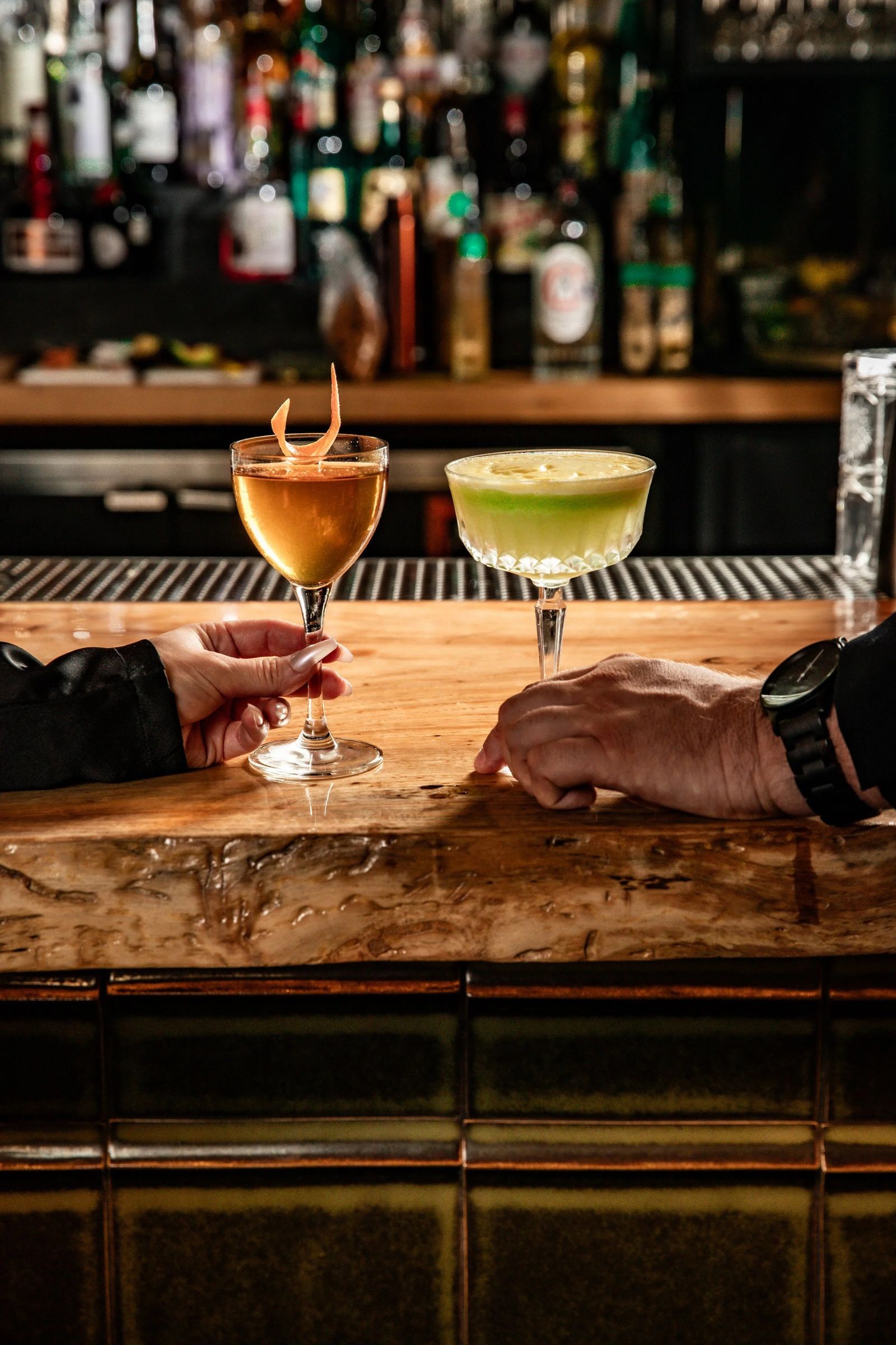 Two hands holding cocktails over a wooden bar with shelves of bottles behind it.