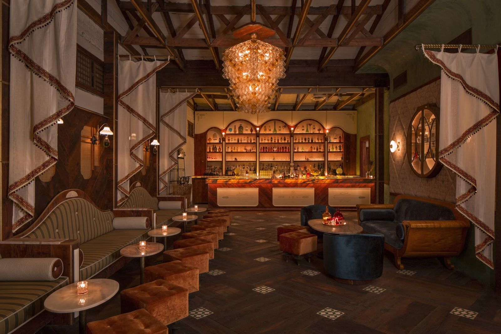 A candlelit bar room with a chandelier, wooden ceiling beams, banquette seating and a lit back bar.