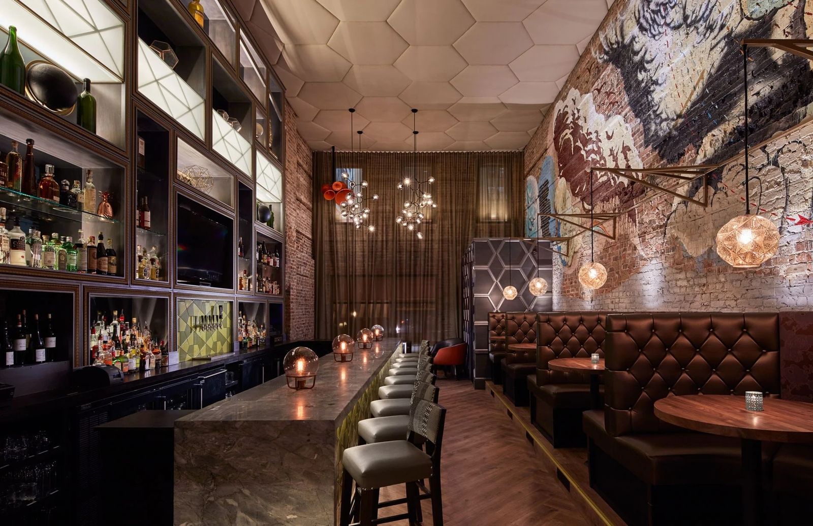 A long bar with leather booths along a brick wall, hanging lamps and a mural with bottles on shelves.