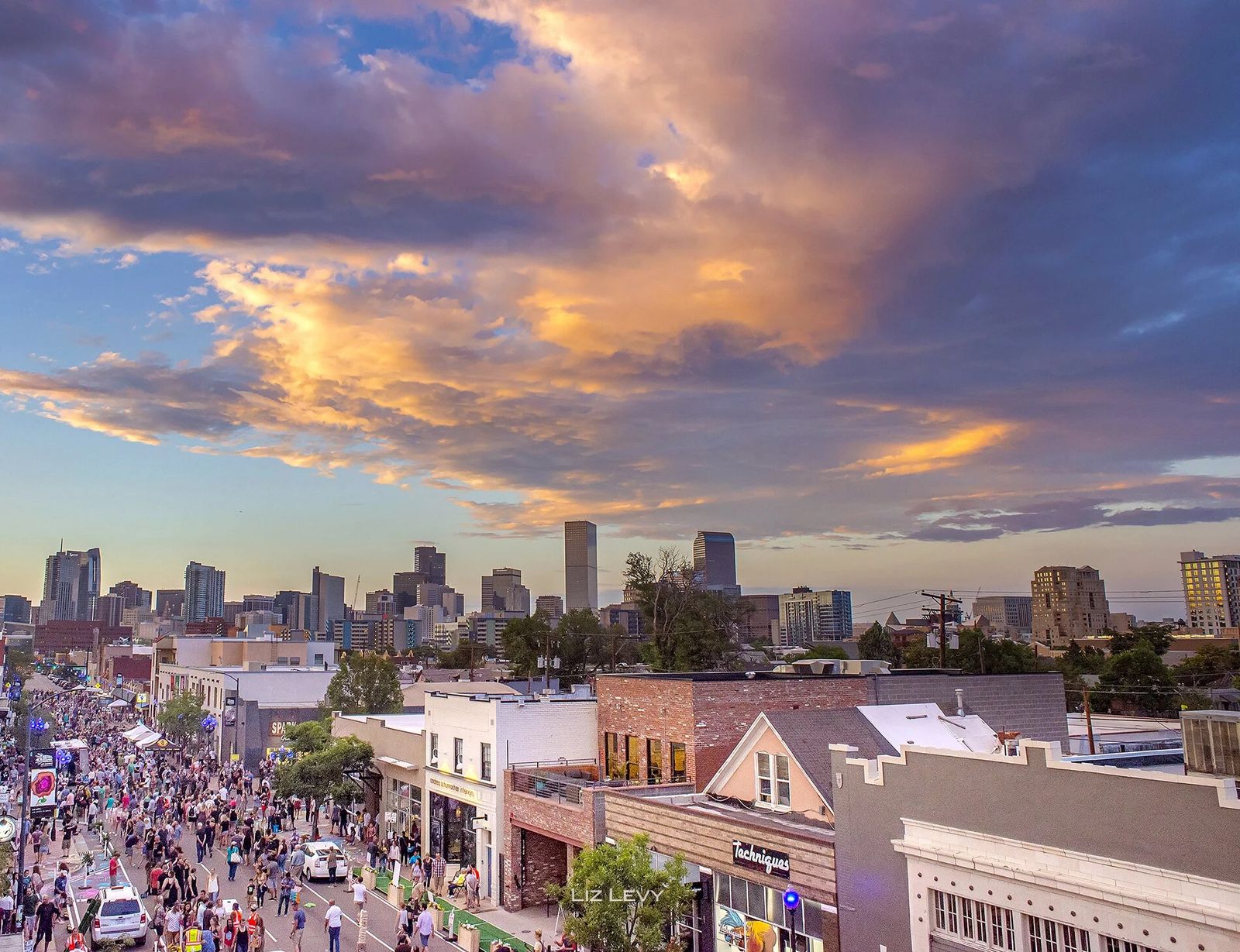 A busy street at sunset with a crowd between low buildings and downtown towers beyond.