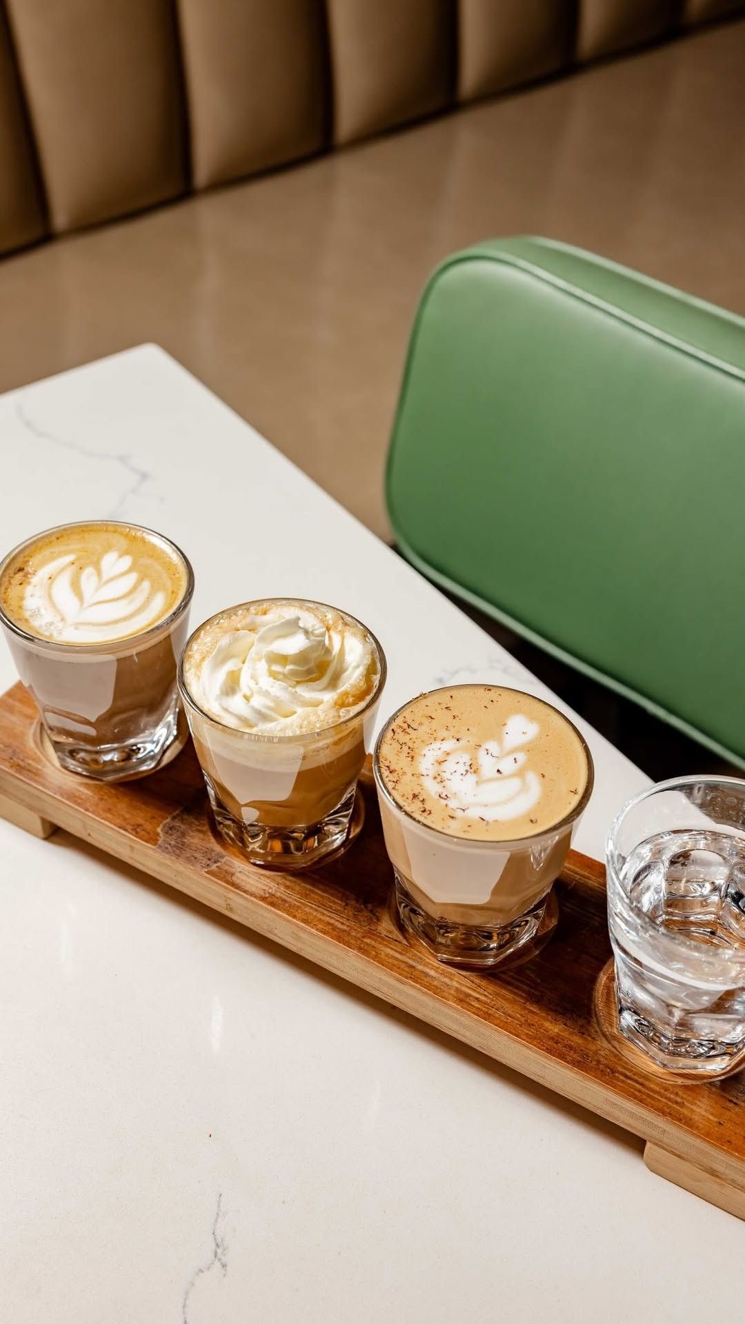A flight of three small lattes on a wooden tray on a white marble counter.