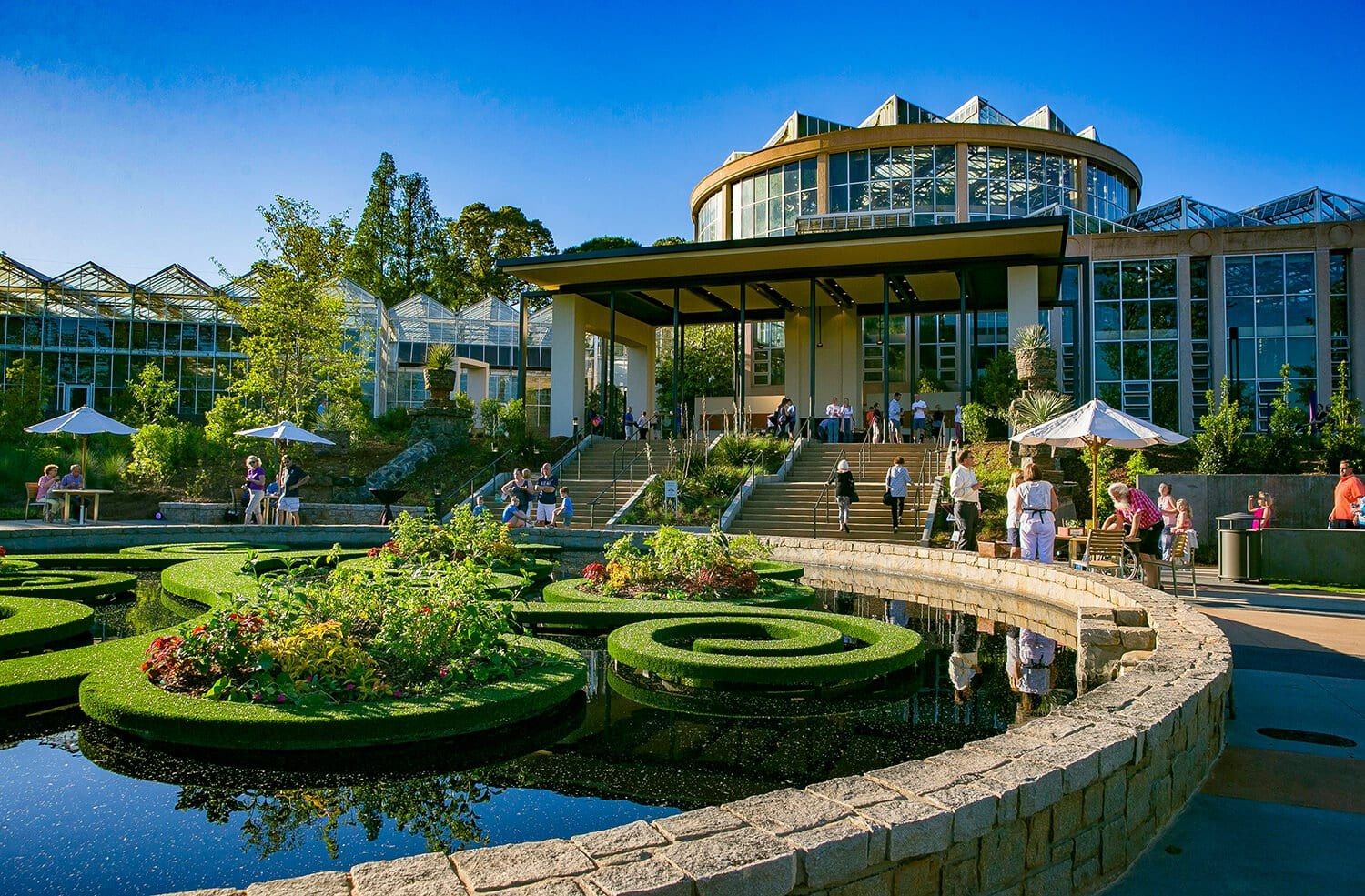A garden pavilion with glass-walled conservatory behind a curved reflecting pool and manicured hedges