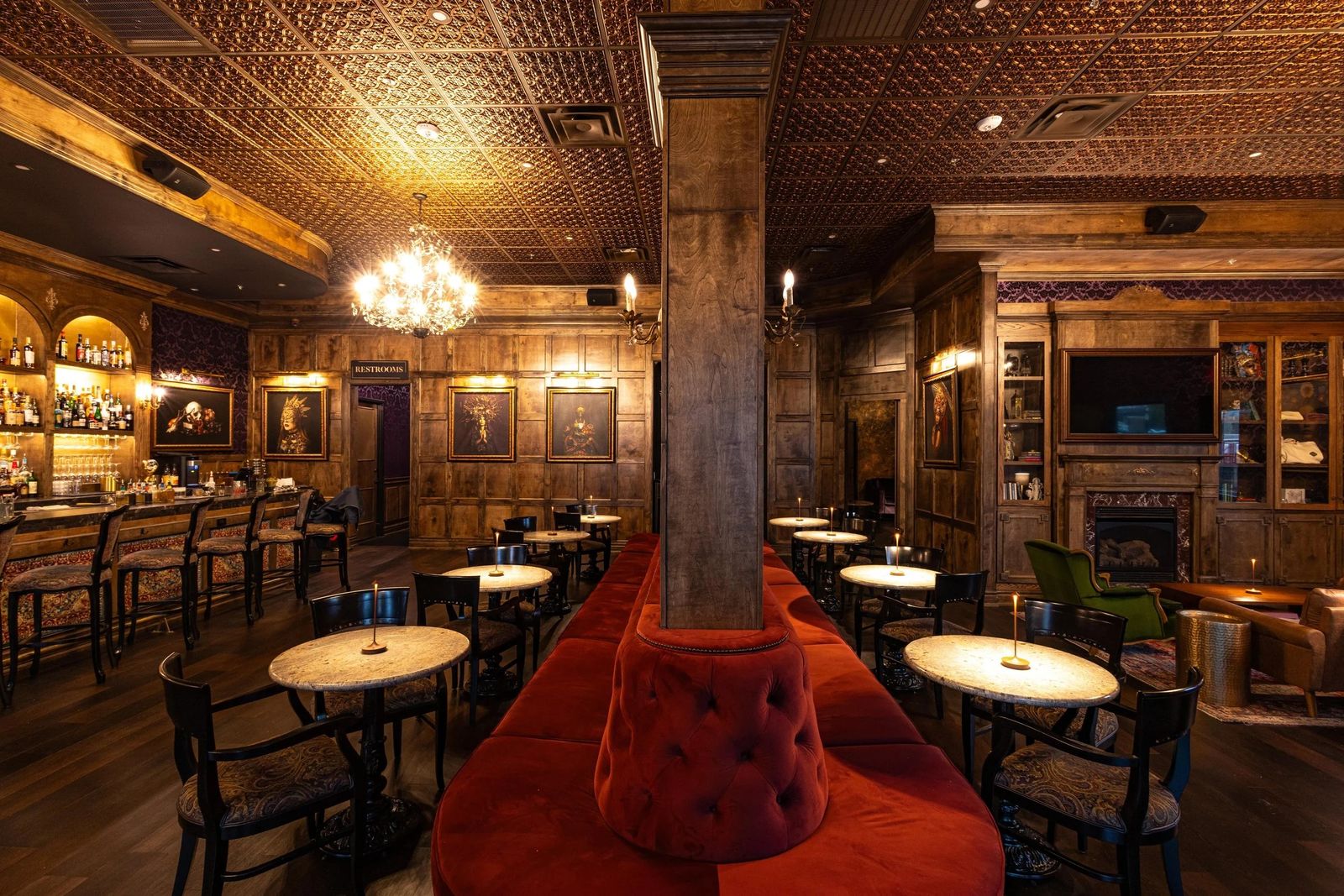 A wood-paneled bar room with a long red banquette, small round tables, chandeliers and a back bar of bottles.