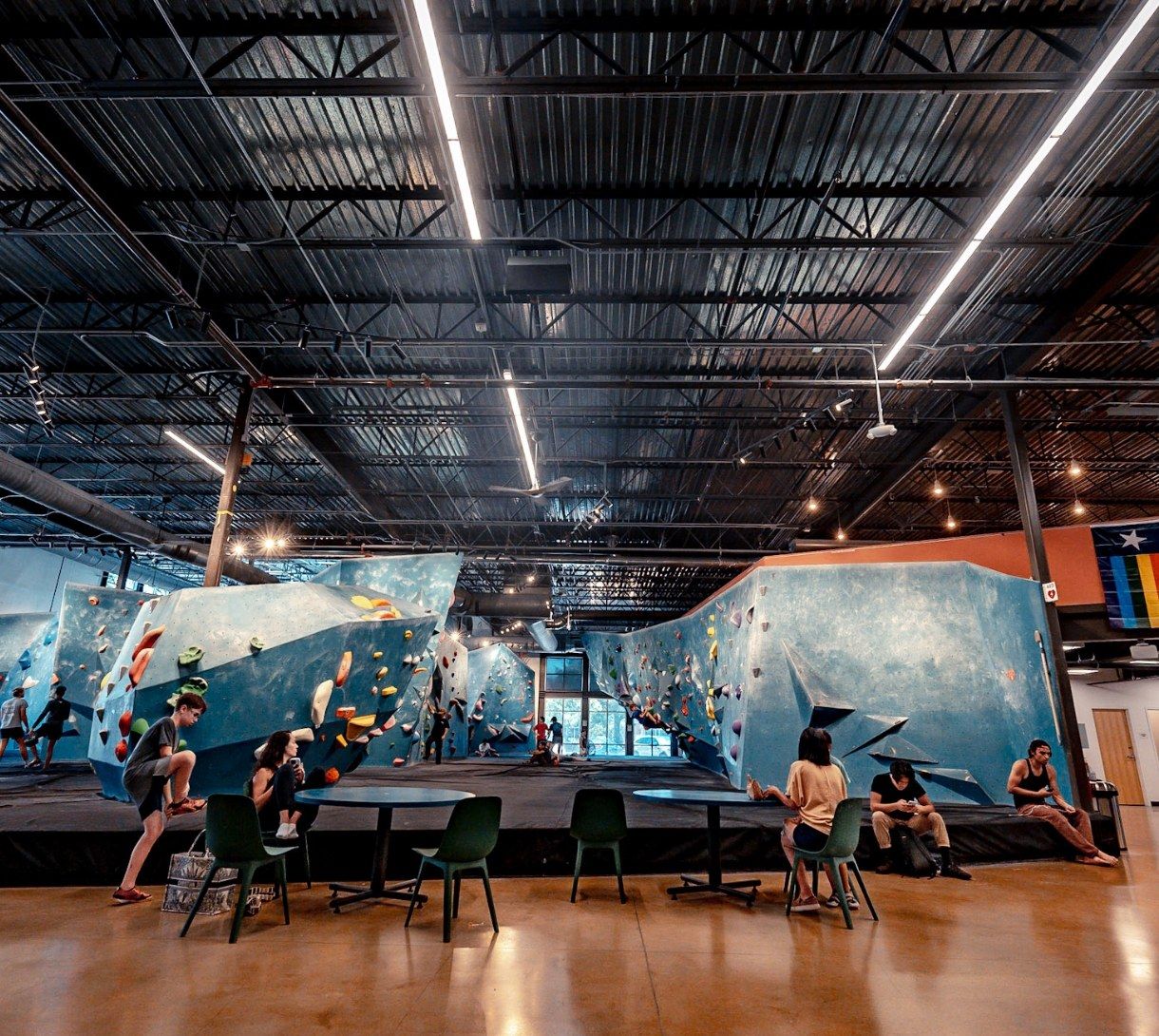 A large bouldering gym with blue climbing walls, a high ceiling with skylights and lounge tables below.
