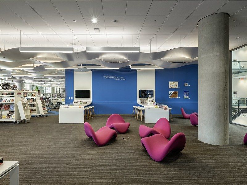 A library floor with a blue wall, concrete columns and rows of bookshelves, with magenta lounge chairs in front.