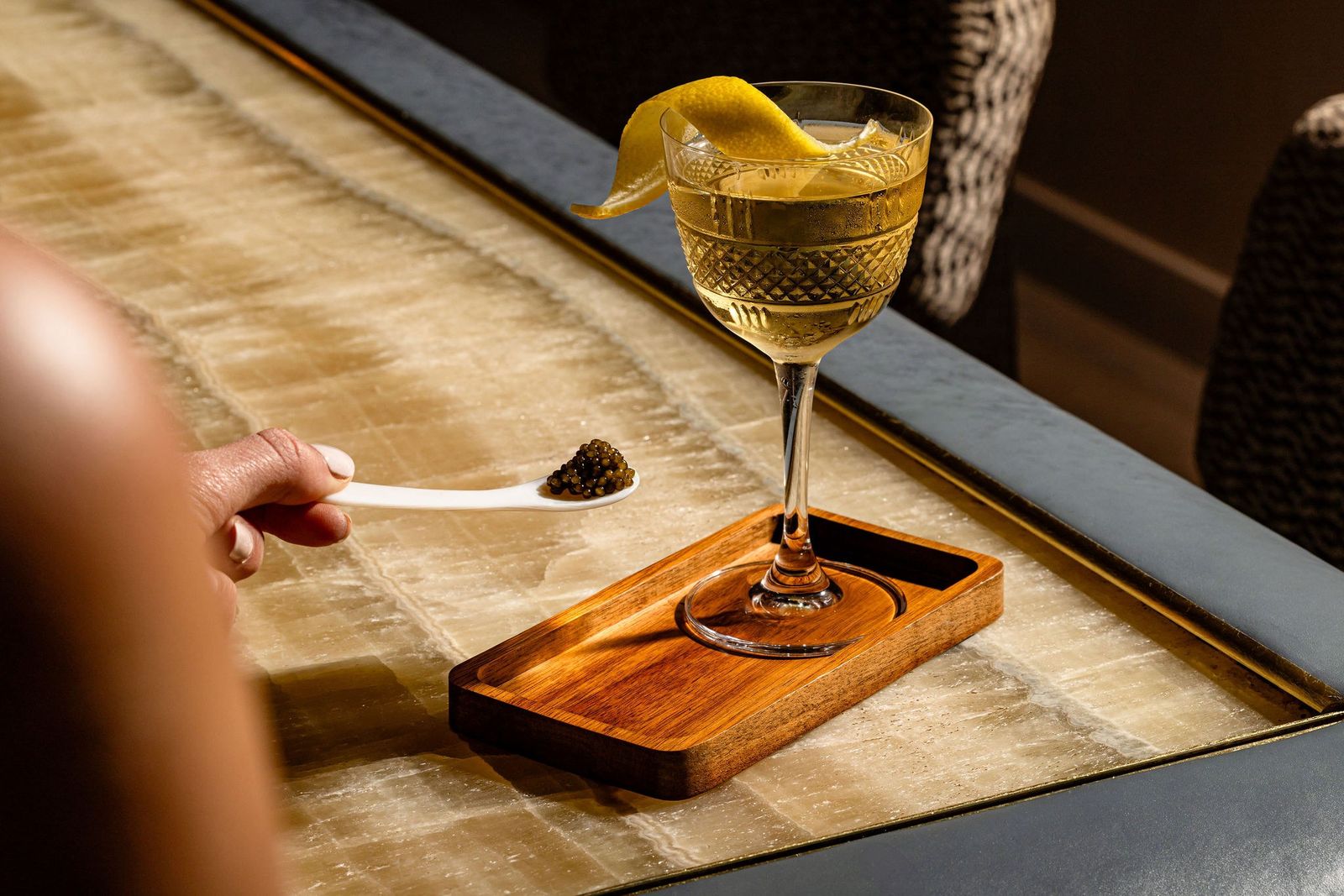 Coupe cocktail with a lemon twist on a wooden tray beside a small spoon of dark garnish