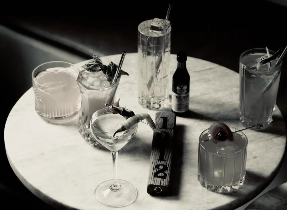 Black-and-white photo of six cocktails on a round marble table beside a wooden 1912 sign
