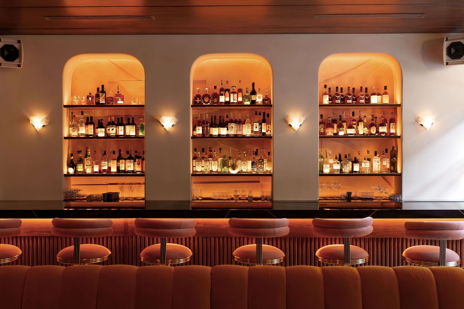 A back bar with three arched, lit shelf niches of bottles above a row of cushioned stools.
