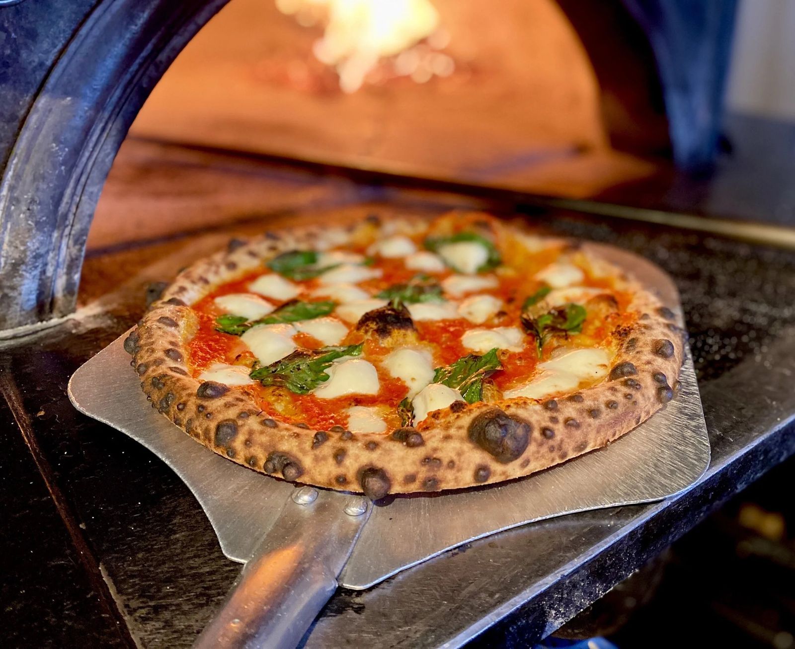 A pizza with basil and mozzarella on a metal peel in front of a wood-fired oven.