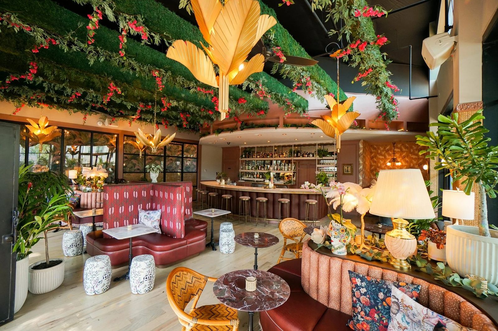 A lounge with pink banquettes, round tables, a central bar and walls and ceilings covered in green plants and orange flowers.