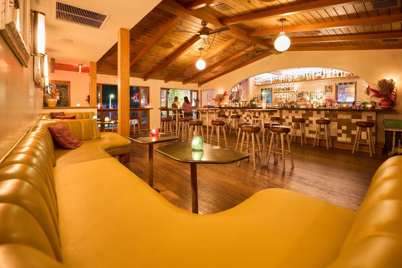 Warmly lit bar room with a mustard-yellow banquette, wood ceiling and a long bar with stools
