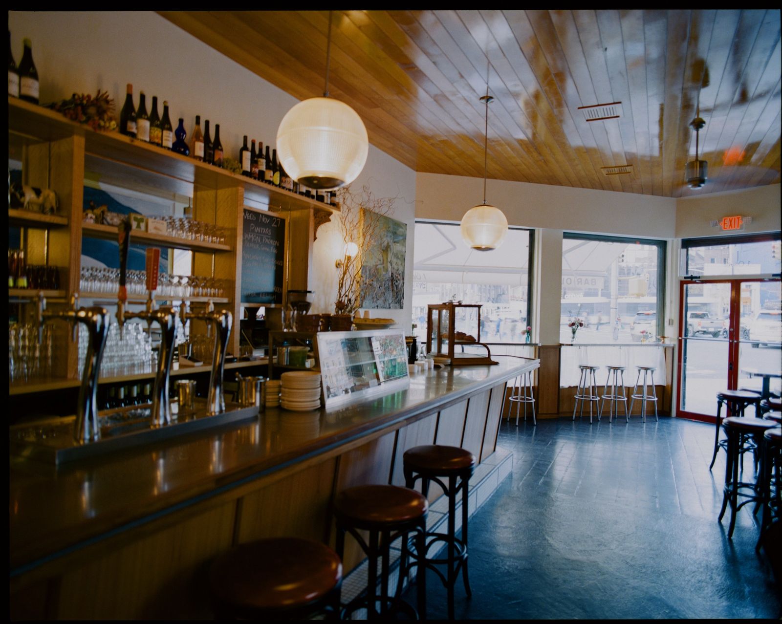 A long wooden bar with taps and bottle shelves in a bright room with large windows