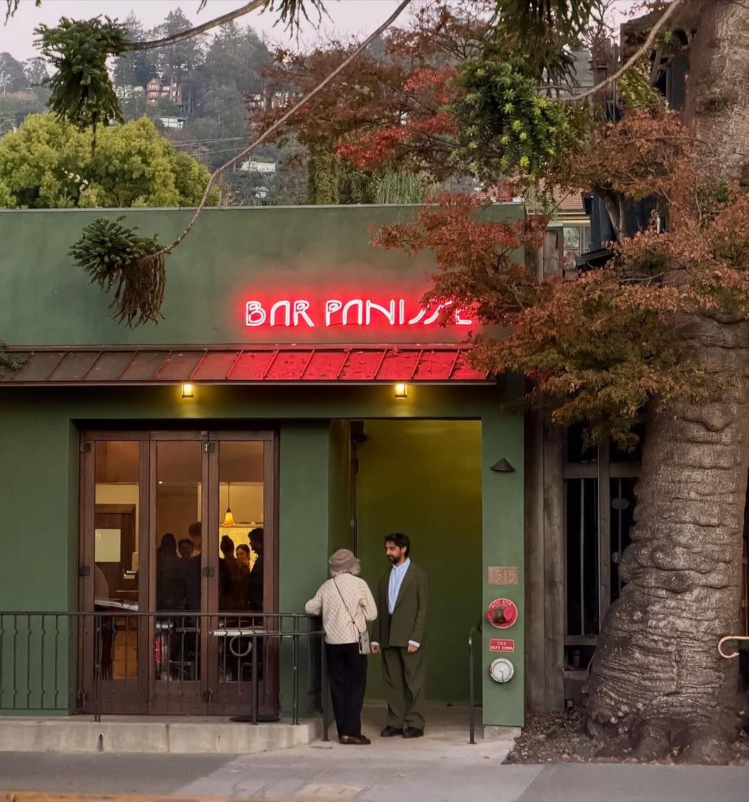 A green storefront with a pink neon Bar Panisse sign and wooden doors, under a large tree.