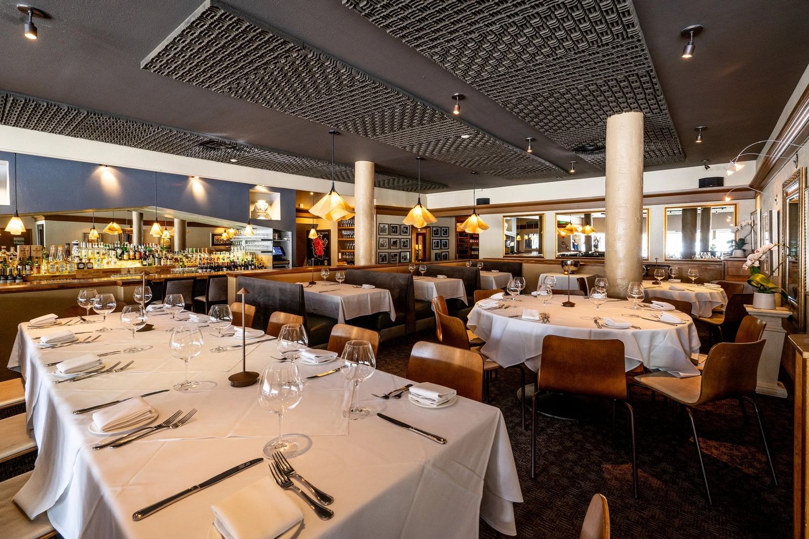 A restaurant dining room with white-clothed tables, tan chairs and a long bar behind, under a patterned ceiling.