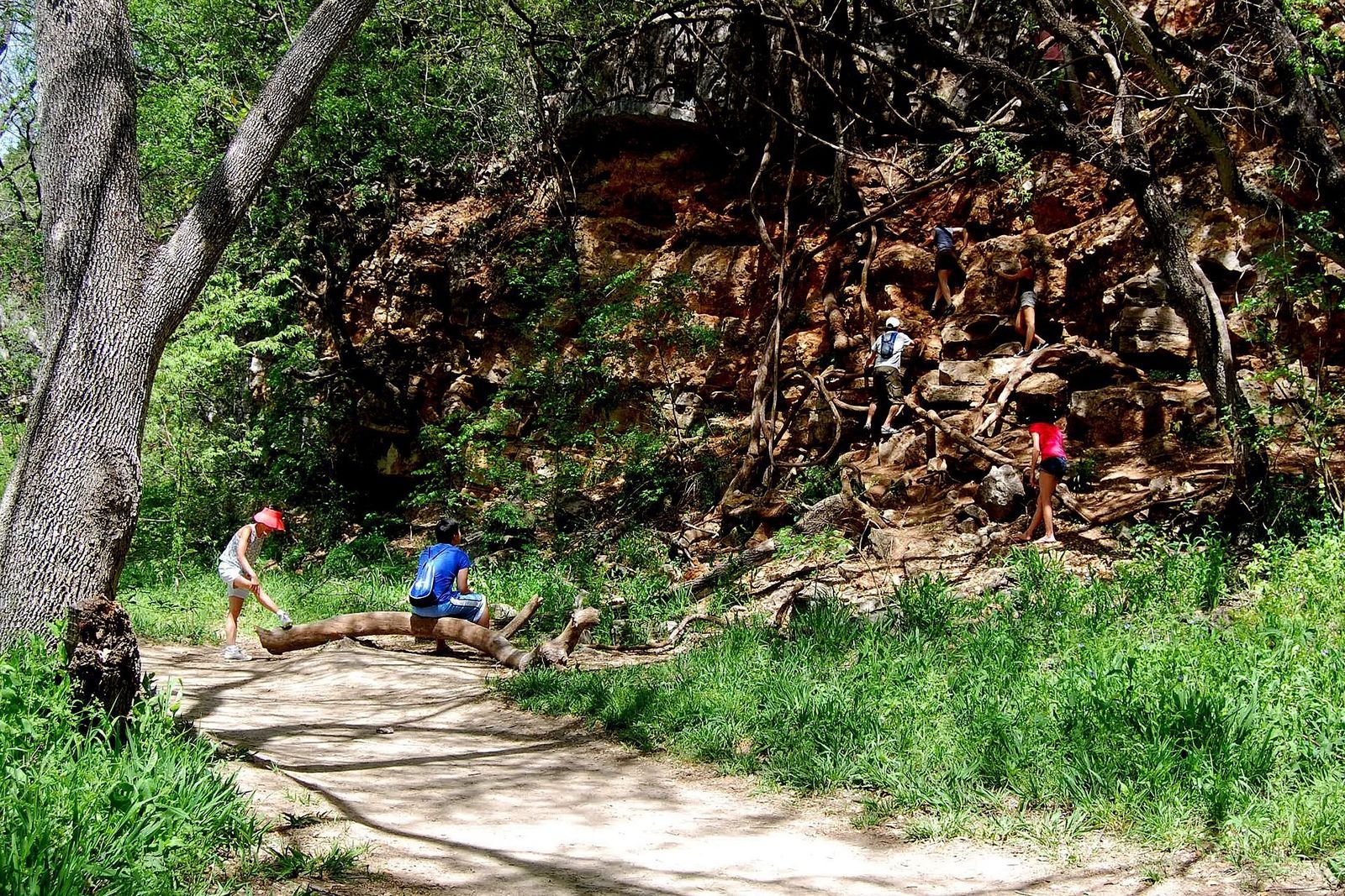 A dirt trail through green trees and grass beside a red rock wall, with a few people in the distance.