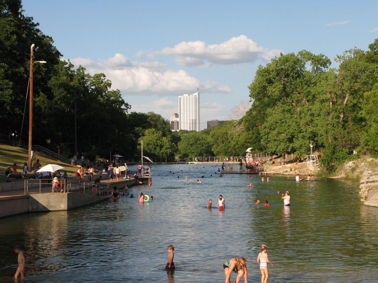 Swimmers in a long spring-fed pool lined with trees, with a few buildings of the city behind.