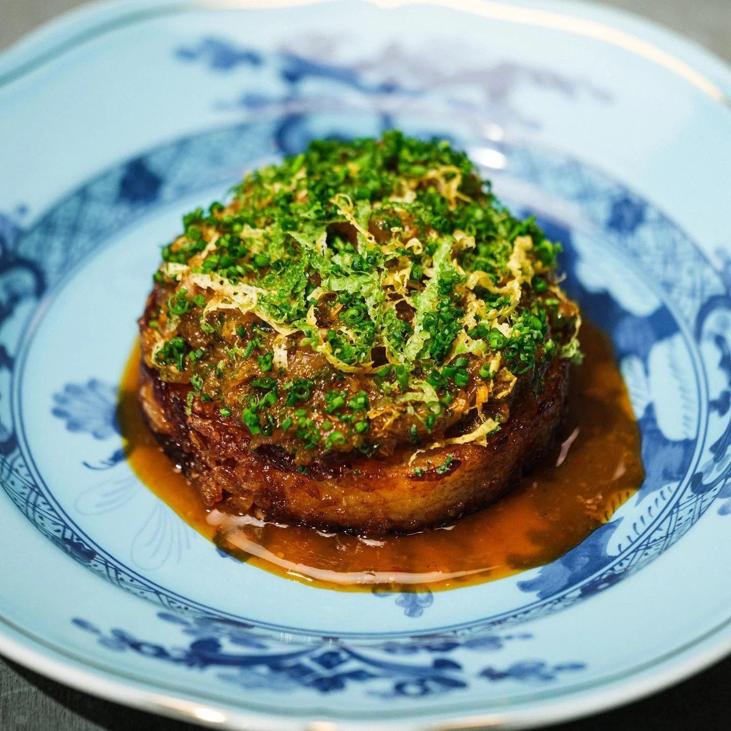 Round crispy fritter topped with shredded herbs in a glossy sauce on a blue and white patterned plate