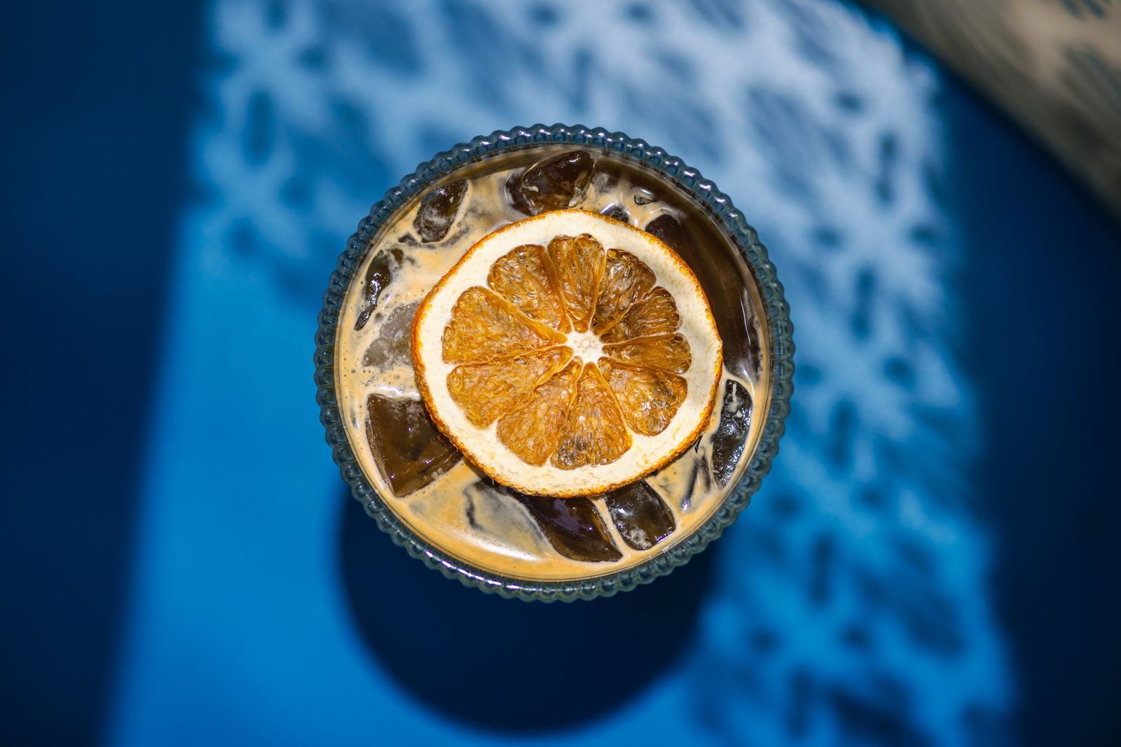 An iced drink with a dried orange wheel on a blue table in dappled light.