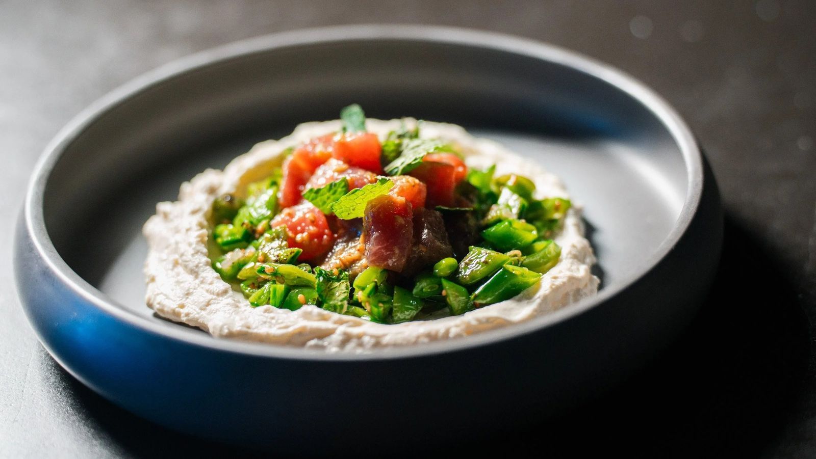 A shallow bowl of whipped white spread topped with chopped green and red vegetables on a dark table.