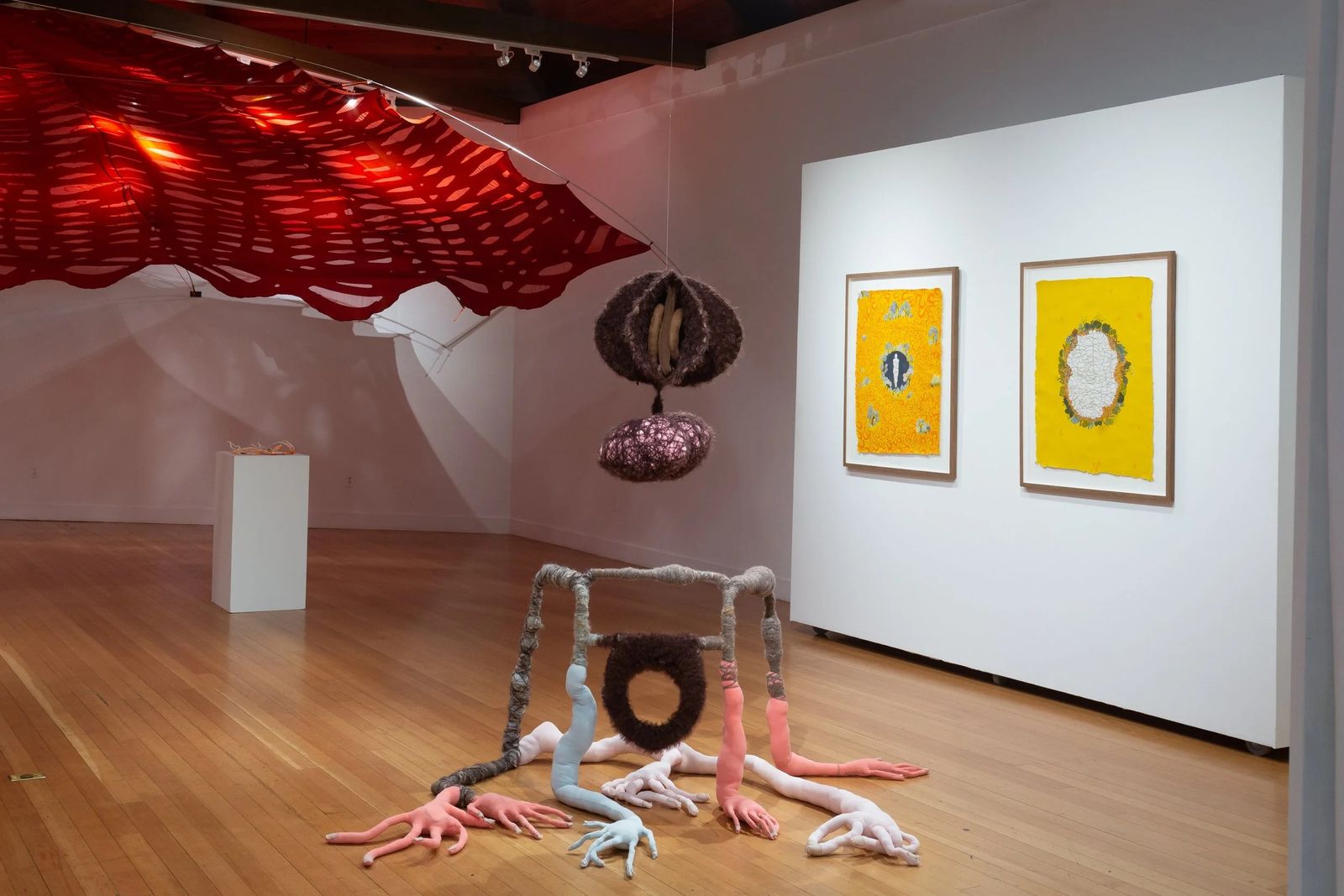 A bright gallery with framed yellow prints on a white wall and a sculpture on the floor under a red canopy.