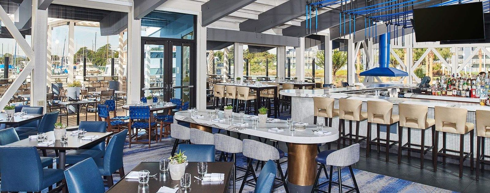 A bright waterfront dining room with blue chairs, white tables, a long bar counter and large windows onto the marina.