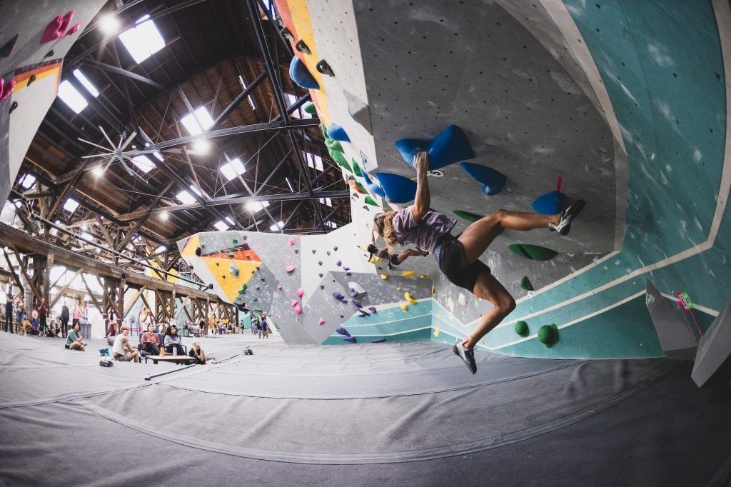 A climber on an angled bouldering wall inside a large warehouse with a high ceiling and skylights.