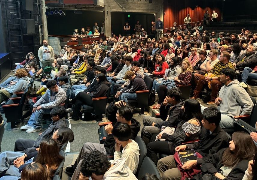 A packed audience seated in rows in a theater house, with some people standing at the back.