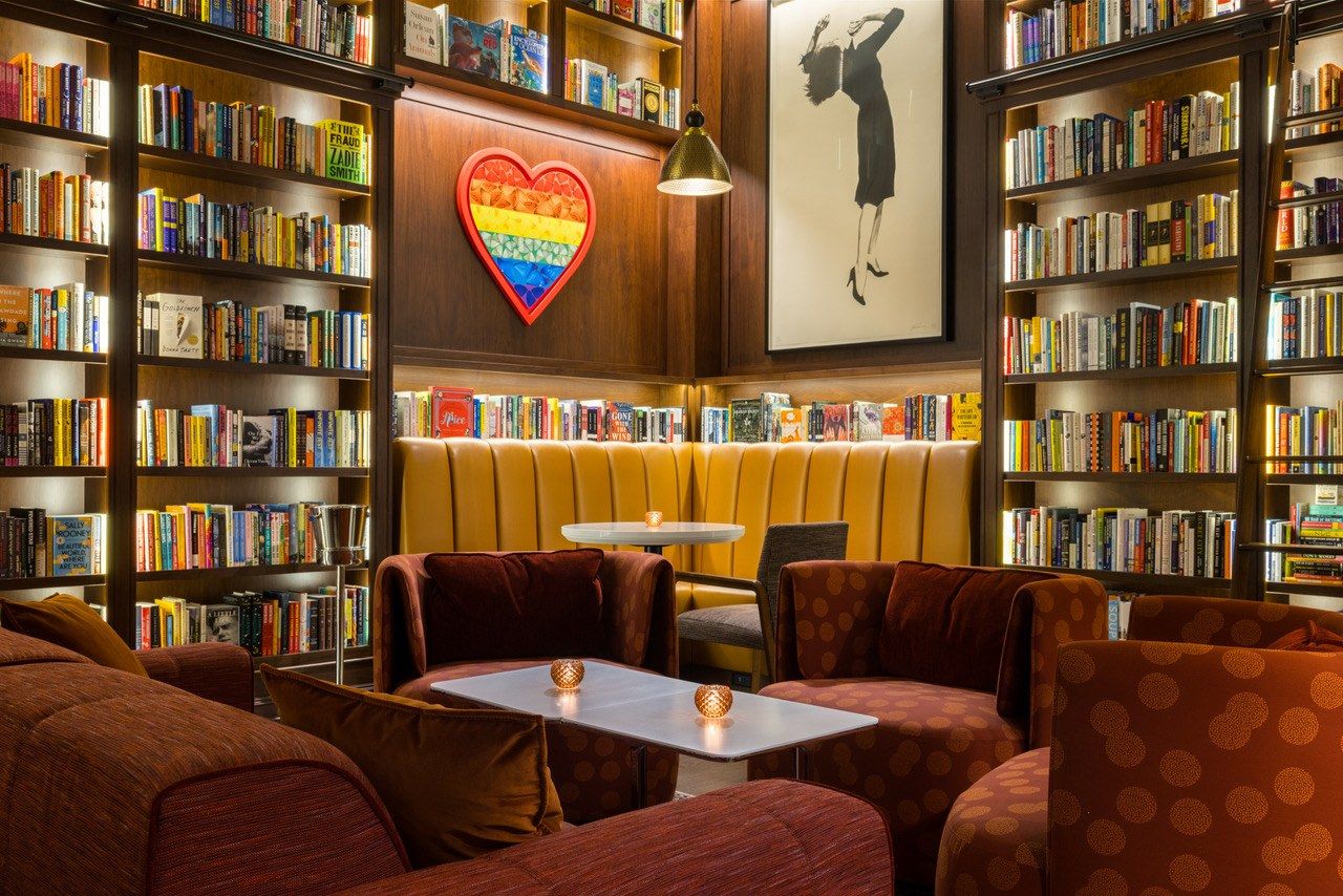A library-style lounge with floor-to-ceiling bookshelves, a yellow banquette and dark red sofas around small tables.