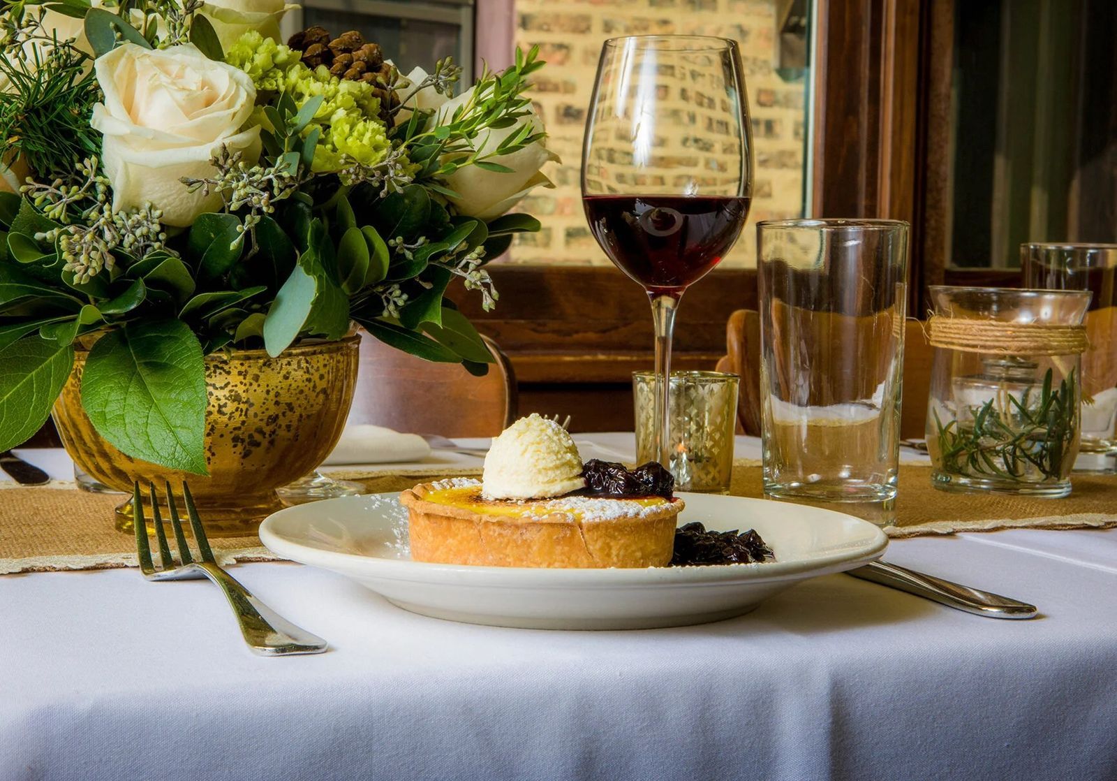 A table with a vase of white roses, a wine glass and a tart on a plate beside a napkin on a white tablecloth.