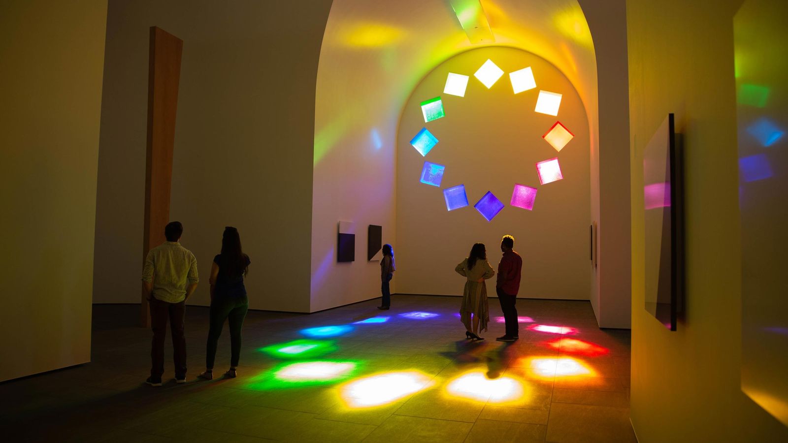 A chapel-like room with arched windows casting colored light patterns onto the floor and walls, with two visitors.