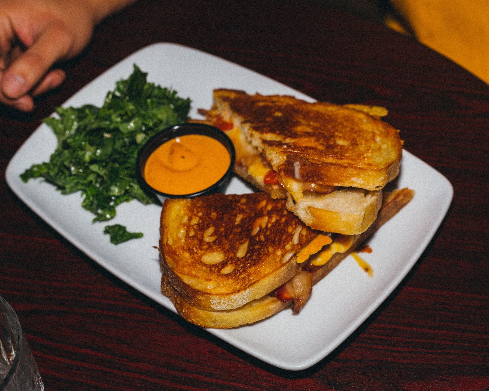 A grilled cheese sandwich cut in half beside a green salad and a small cup of orange sauce on a white plate.