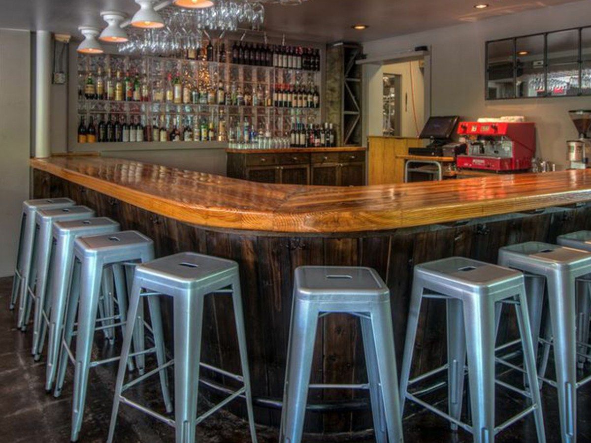 Wooden bar counter lined with metal stools in front of a shelf of bottles