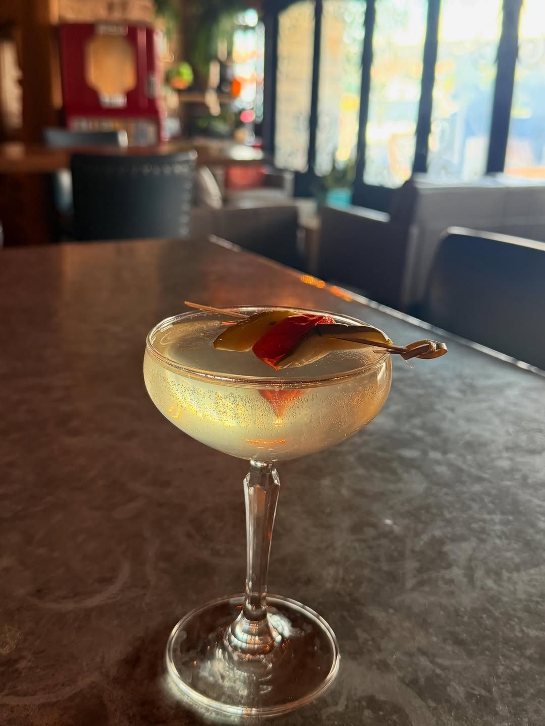 A coupe cocktail with a twist and a small garnish on a dark bar top, with bottles blurred behind.