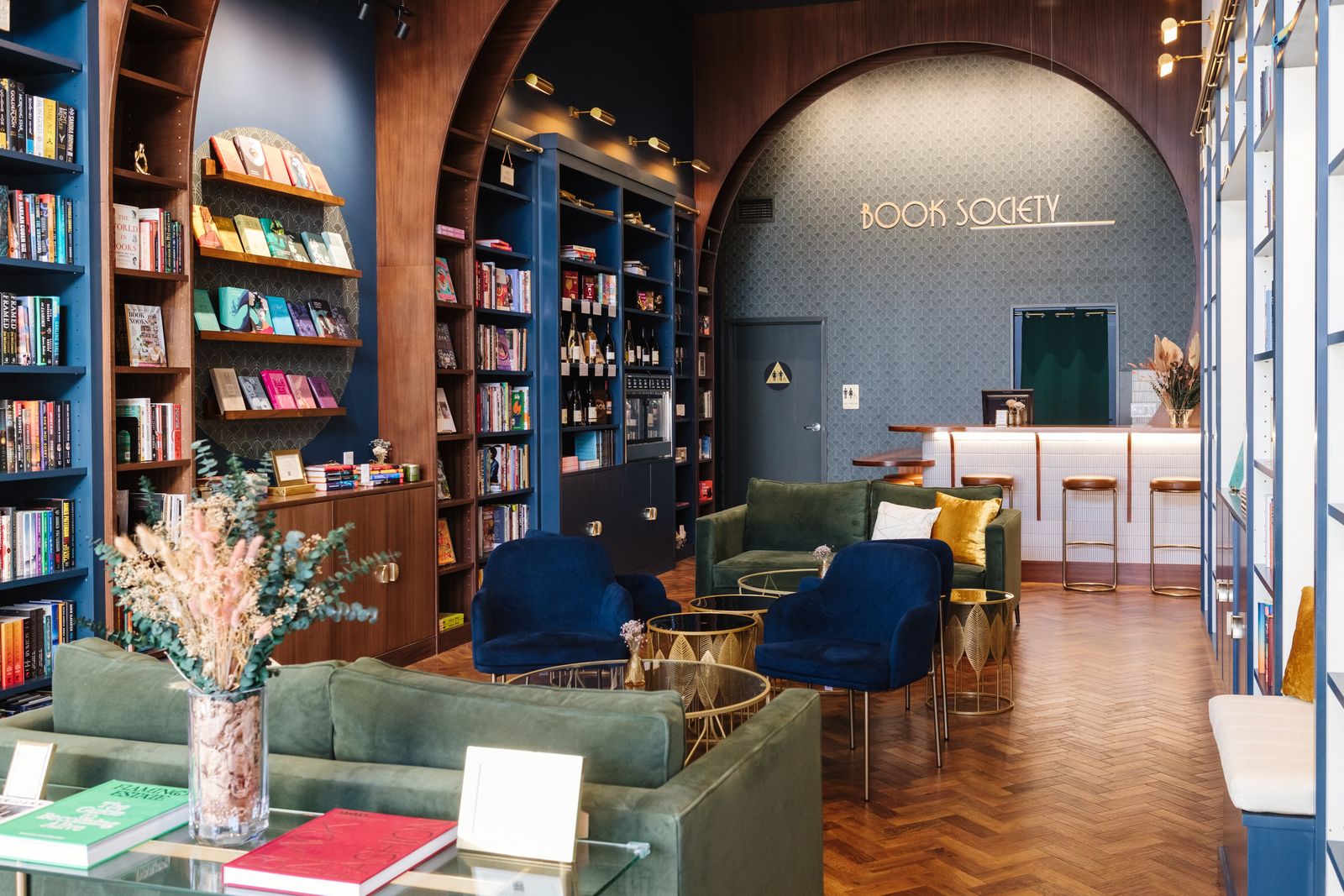 A bookstore lounge with tall shelves, green sofas and navy armchairs, and a bar counter under a dark arch that reads Book Society.