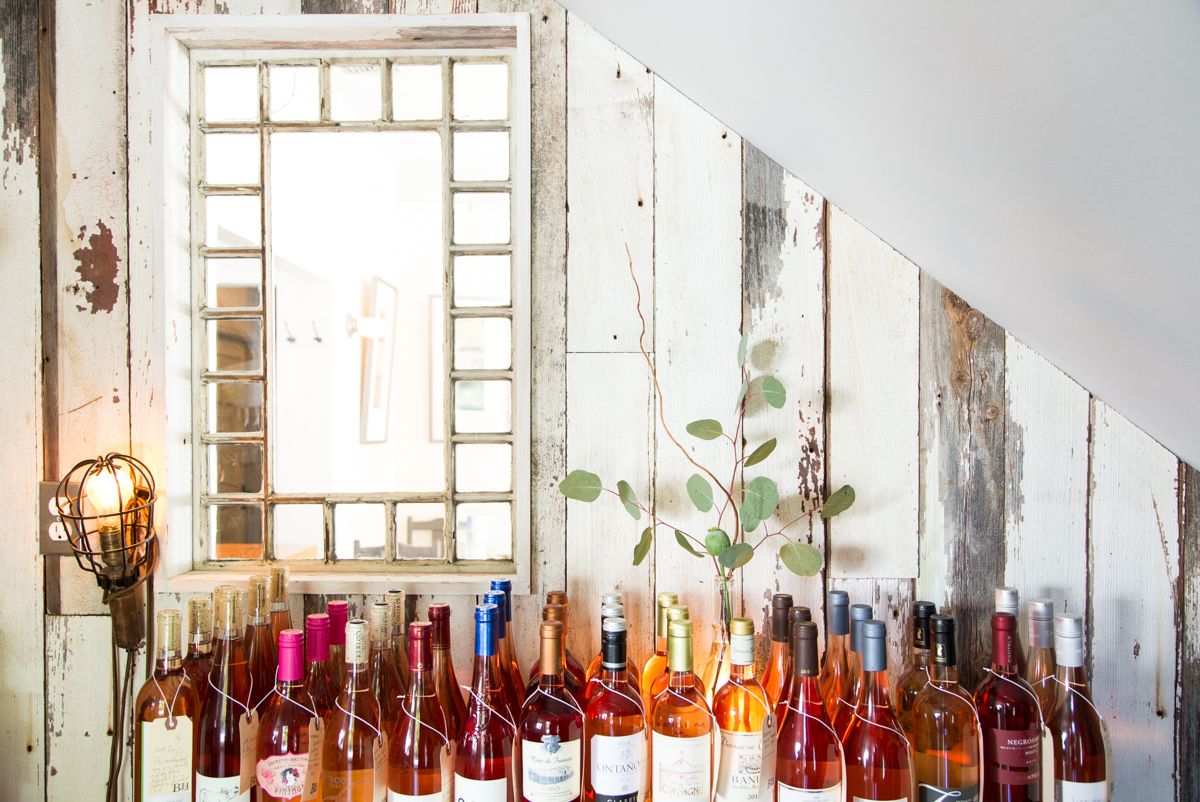 A shelf of wine bottles in front of a whitewashed plank wall with a mirror and a small lamp.