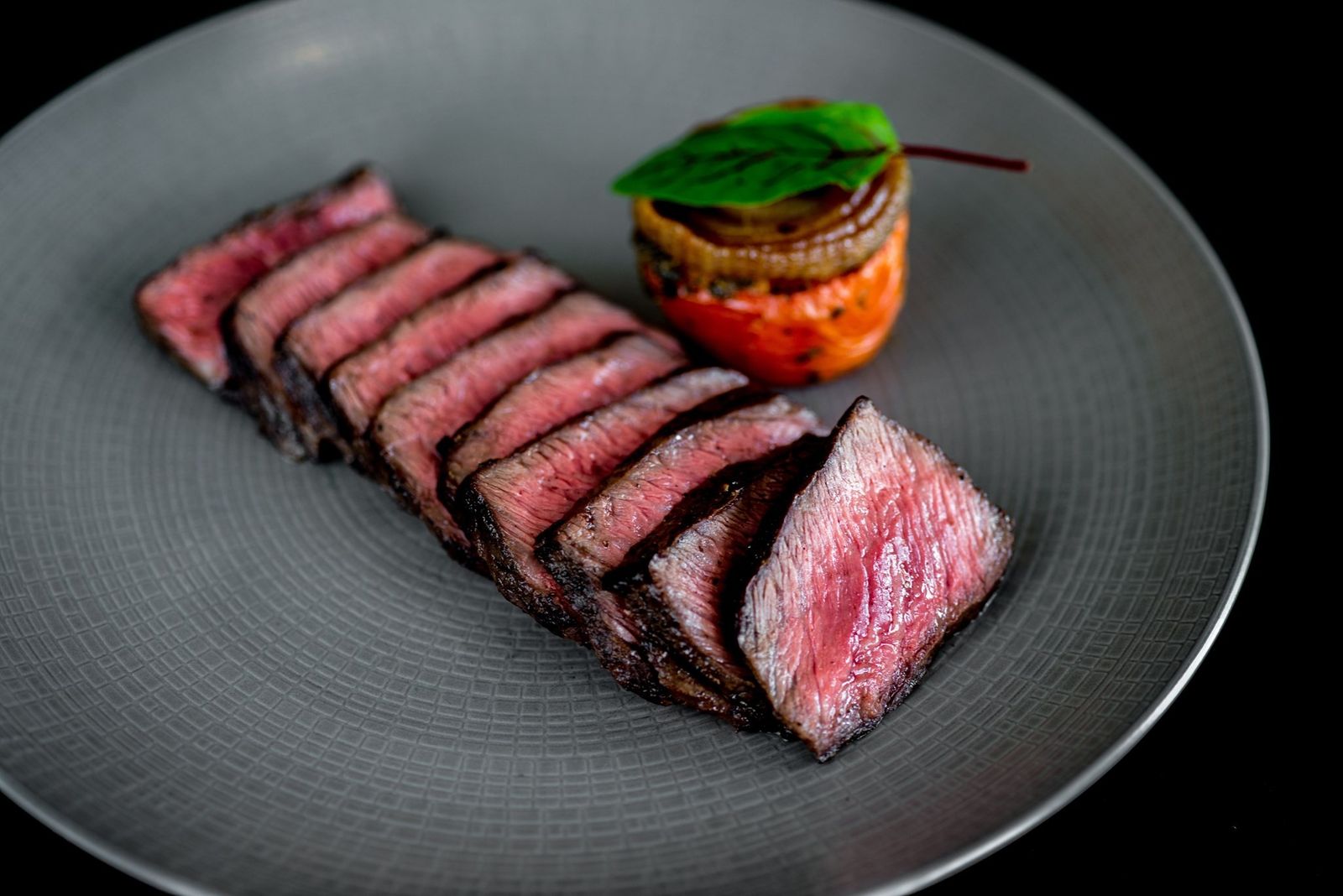 Sliced medium-rare steak fanned on a gray plate beside a small stack of roasted vegetables.