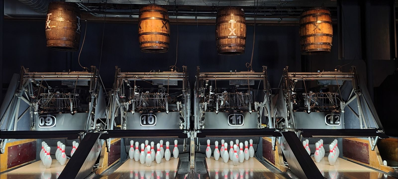 A row of lit bowling lanes with pins and wooden barrel-shaped lamps hanging overhead.