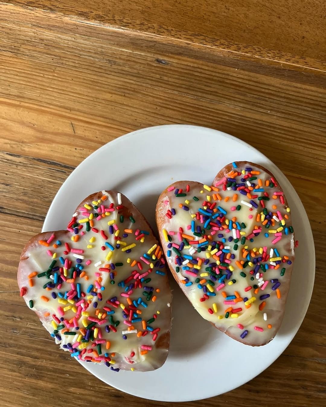 Two heart-shaped donuts with rainbow sprinkles on a white plate on a wooden table.
