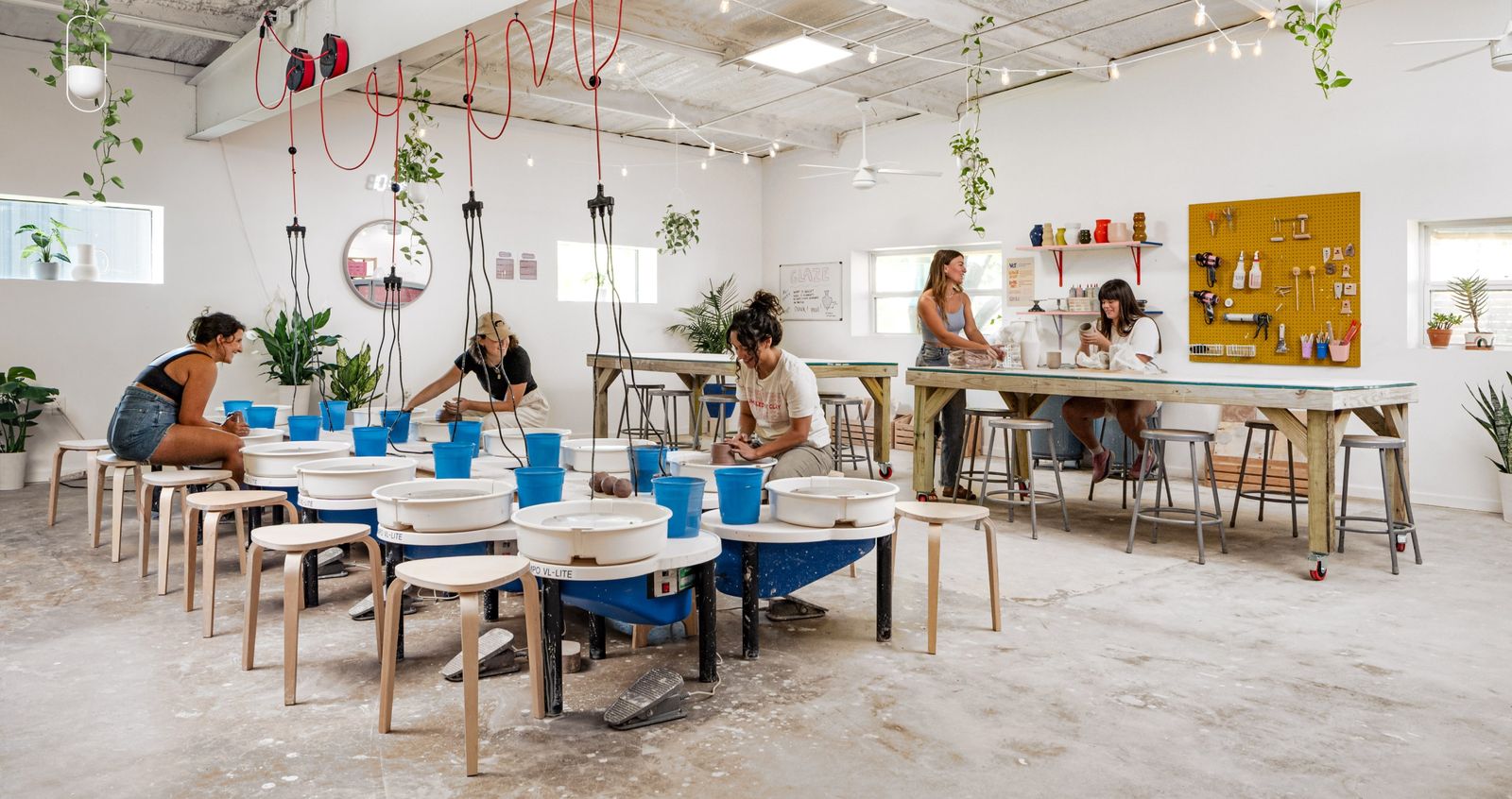 A bright ceramics studio with long tables, pottery wheels and hanging plants.
