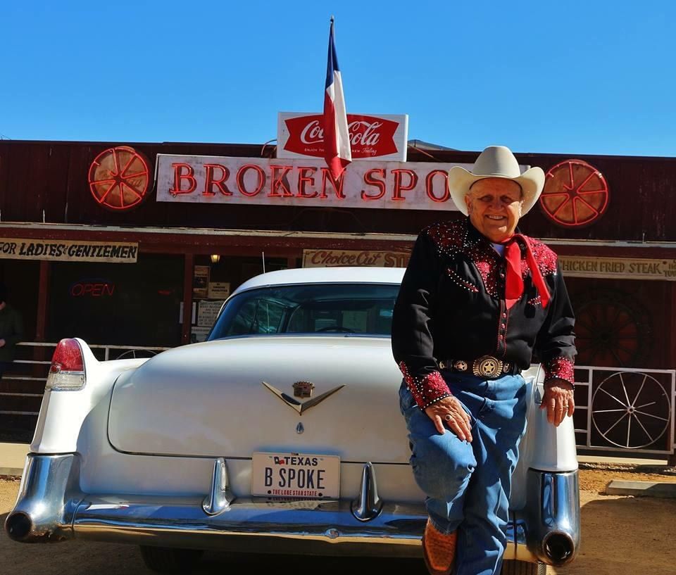 A man in a cowboy hat leaning on a vintage car in front of the Broken Spoke's red building and sign.